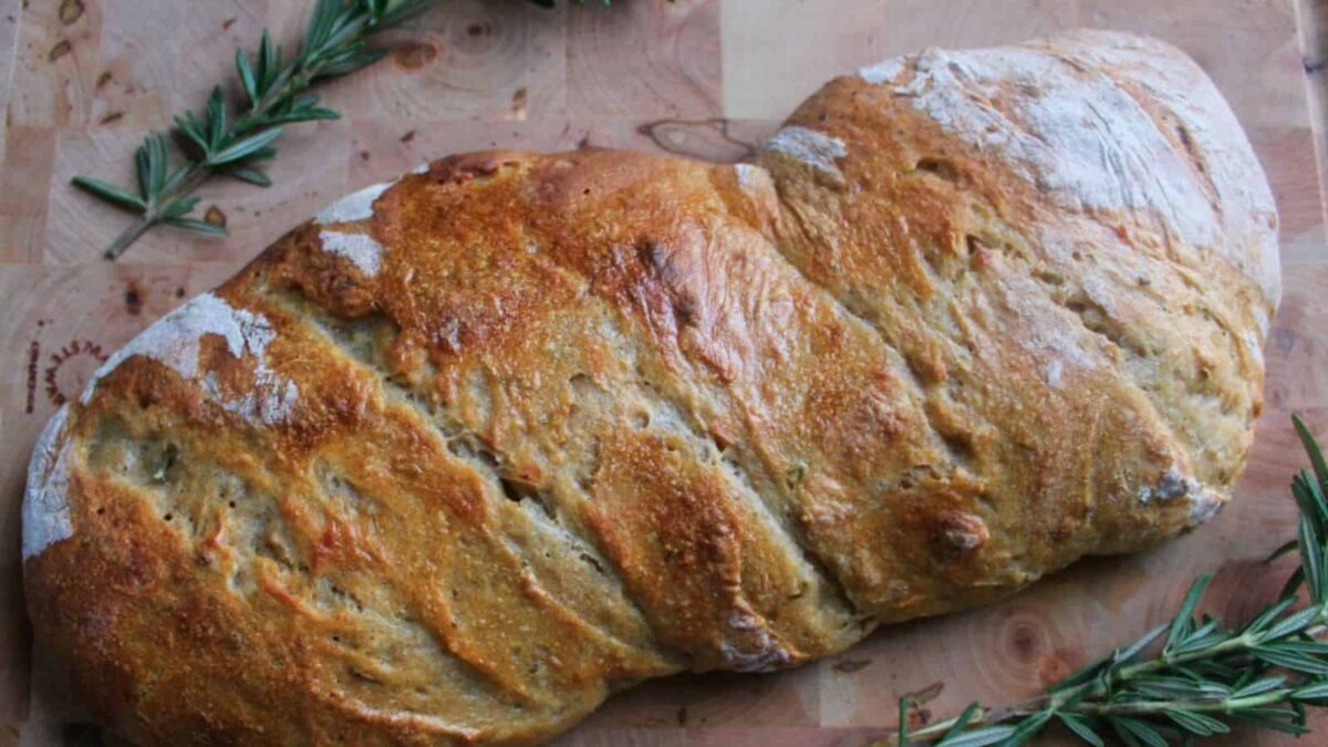 A rustic, golden-brown loaf with a twisted shape rests on a wooden cutting board, surrounded by fresh rosemary, showcasing the rich fermented flavor found in classic sourdough recipes.