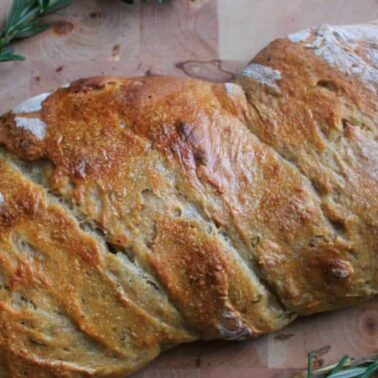 A rustic, golden-brown loaf with a twisted shape rests on a wooden cutting board, surrounded by fresh rosemary, showcasing the rich fermented flavor found in classic sourdough recipes.