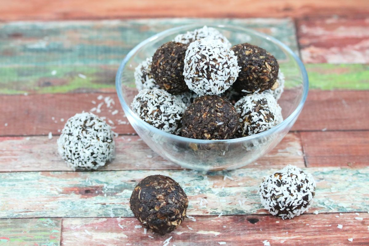 A glass bowl filled with chocolate energy balls, some coated in shredded coconut, sits on a rustic wooden surface. Three balls rest outside the bowl—perfect for fans of coconut desserts.