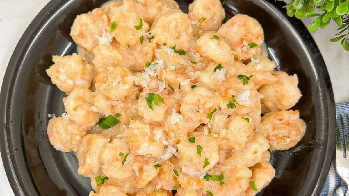 A black plate filled with creamy shrimp, complemented by sweet-savory notes of coconut, garnished with chopped parsley and grated cheese, sits on a white surface next to a small potted plant.