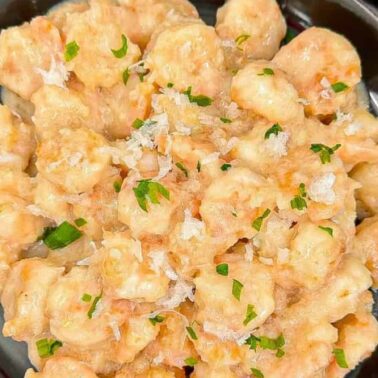 A black plate filled with creamy shrimp, complemented by sweet-savory notes of coconut, garnished with chopped parsley and grated cheese, sits on a white surface next to a small potted plant.