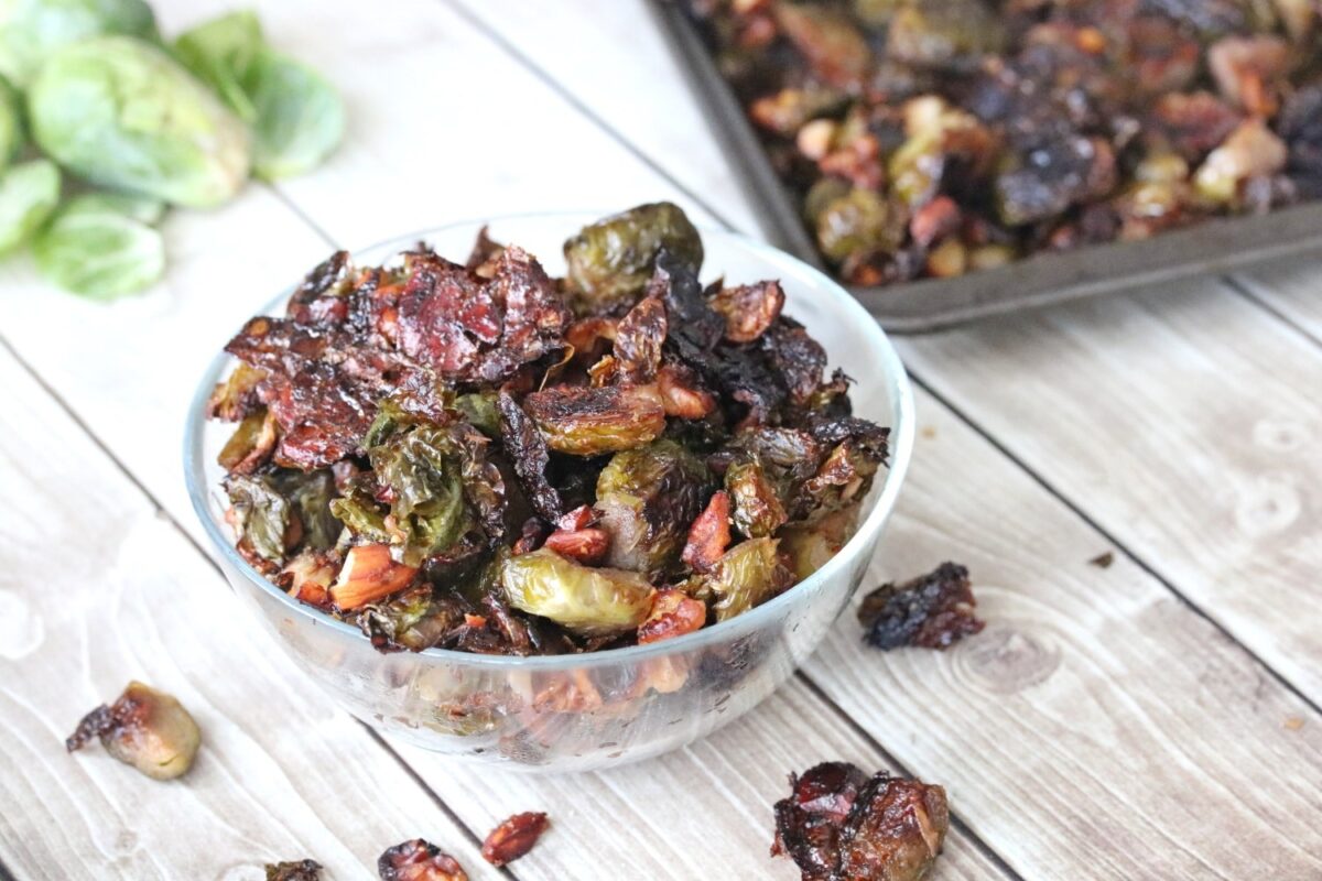 A glass bowl filled with roasted Brussels sprouts and pecans sits on a wooden surface, showcasing a classic fall flavor, with more on a baking tray in the background.