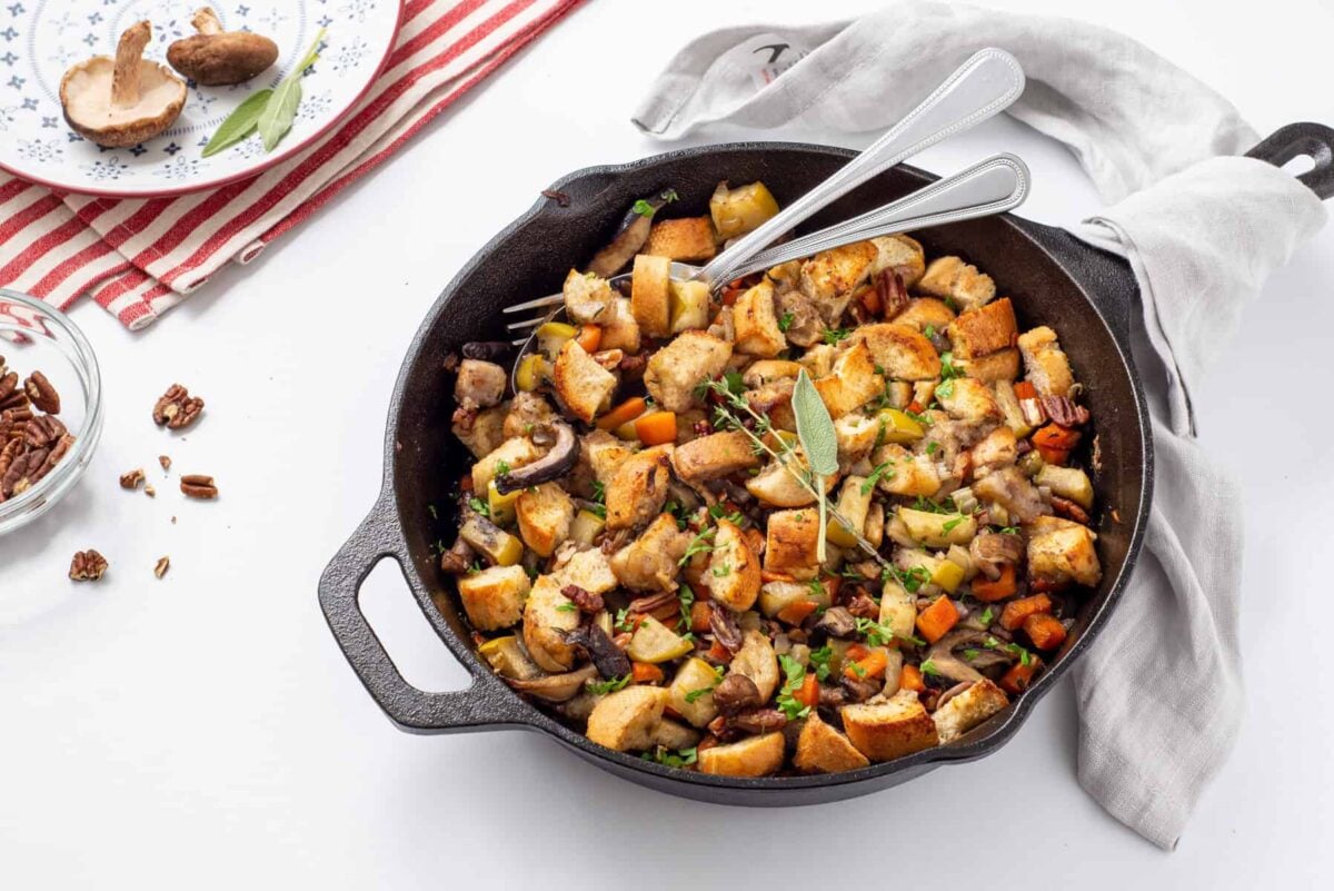A cast iron skillet filled with mixed bread stuffing, vegetables, and herbs—perfect for Thanksgiving stuffing recipes—with two forks inside and a dish towel wrapped around the handle.