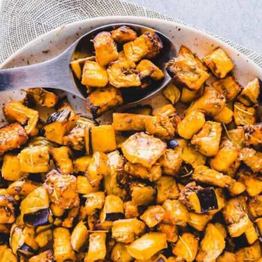 A bowl of roasted, cubed eggplant with herbs, served with a wooden spoon on a neutral-colored cloth—perfect for those seeking flavorful air fryer recipes or easy side dish recipes.