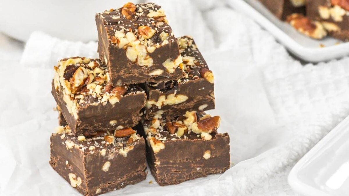 Five pieces of chocolate fudge with chopped pecans are stacked on white parchment paper, with more fudge pieces visible on a plate in the background—a perfect treat for fall baking.