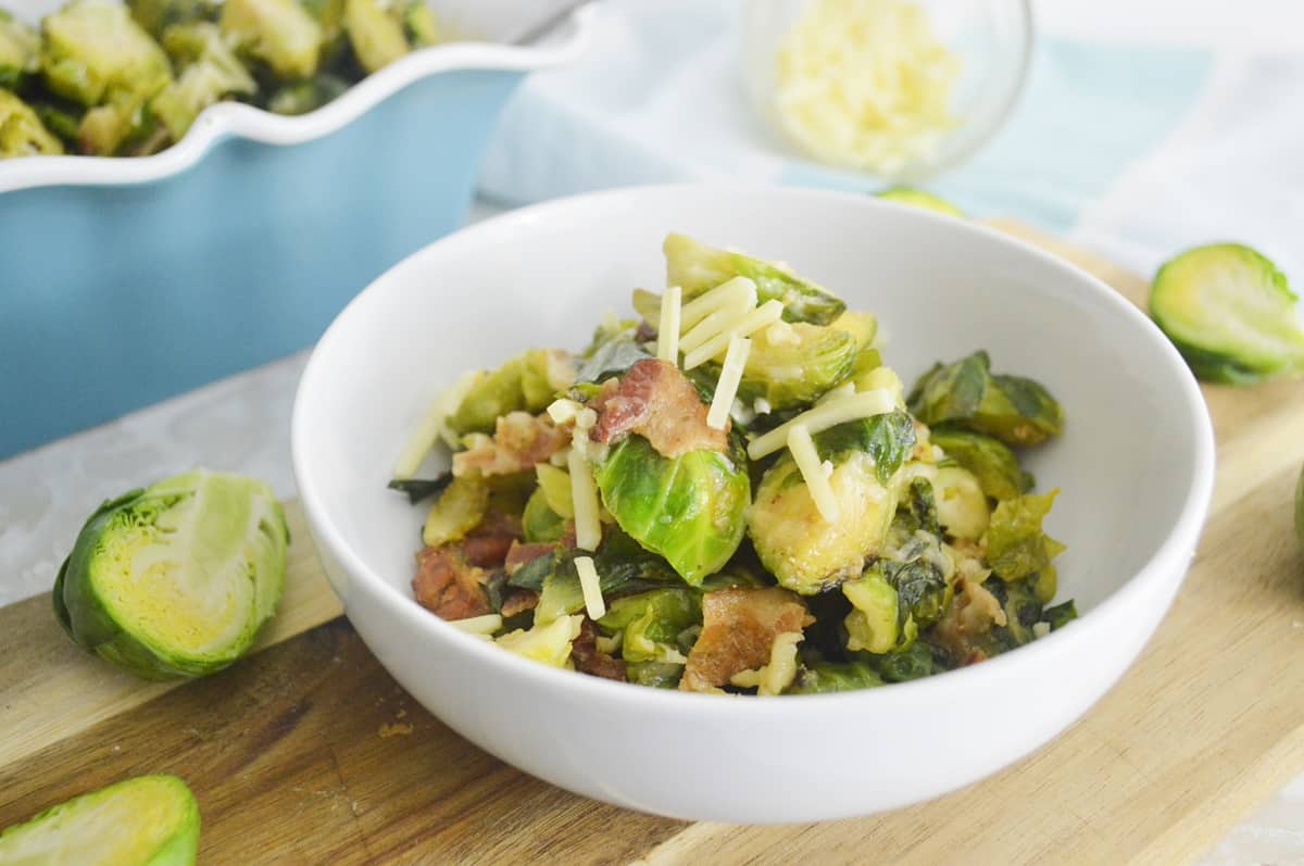 A white bowl filled with Roasted Brussels Sprouts, bacon pieces, and shredded cheese sits on a wooden board with halved Brussels sprouts nearby—perfect for trying new Brussels Sprouts recipes.