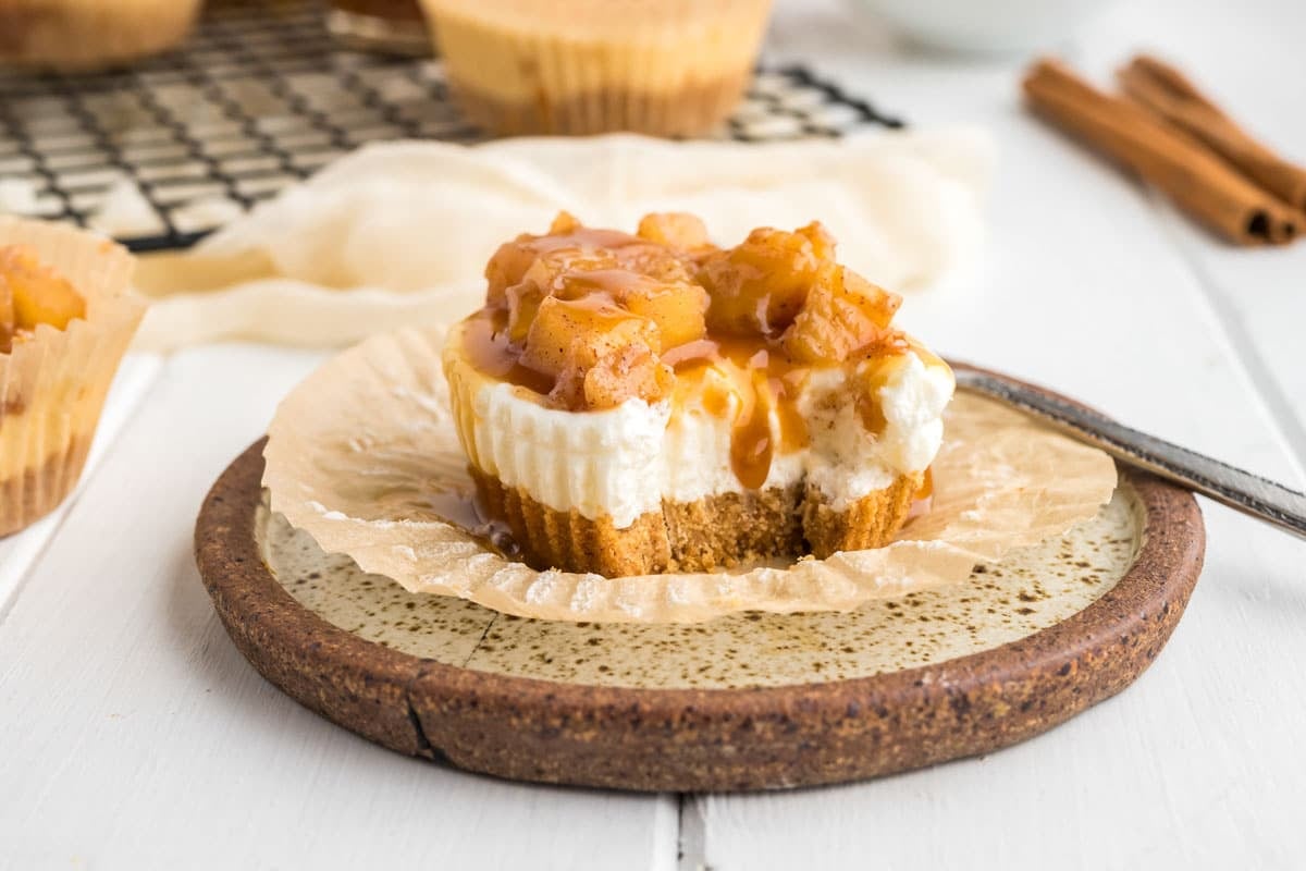 Mini cheesecake topped with caramel sauce and diced cinnamon apples sits on a plate with a bite taken out, next to a fork—an irresistible addition to your favorite Apple desserts, perfect for serving as clean cuts or full slices.