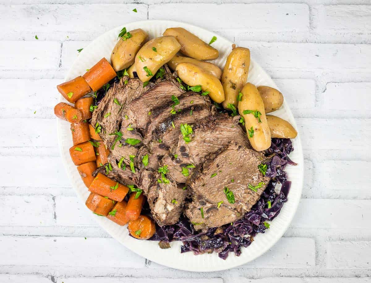 A plate with sliced pot roast, fingerling potatoes, carrots, and shredded red cabbage, garnished with chopped herbs—perfect for those who love hearty Slow Cooker Dinner Recipes—set against a white brick background.
