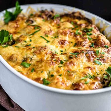 A baked casserole in a white oval dish topped with melted, browned cheese to satisfy your cravings and garnished with fresh parsley—a must-try for fans of cheesy recipes.