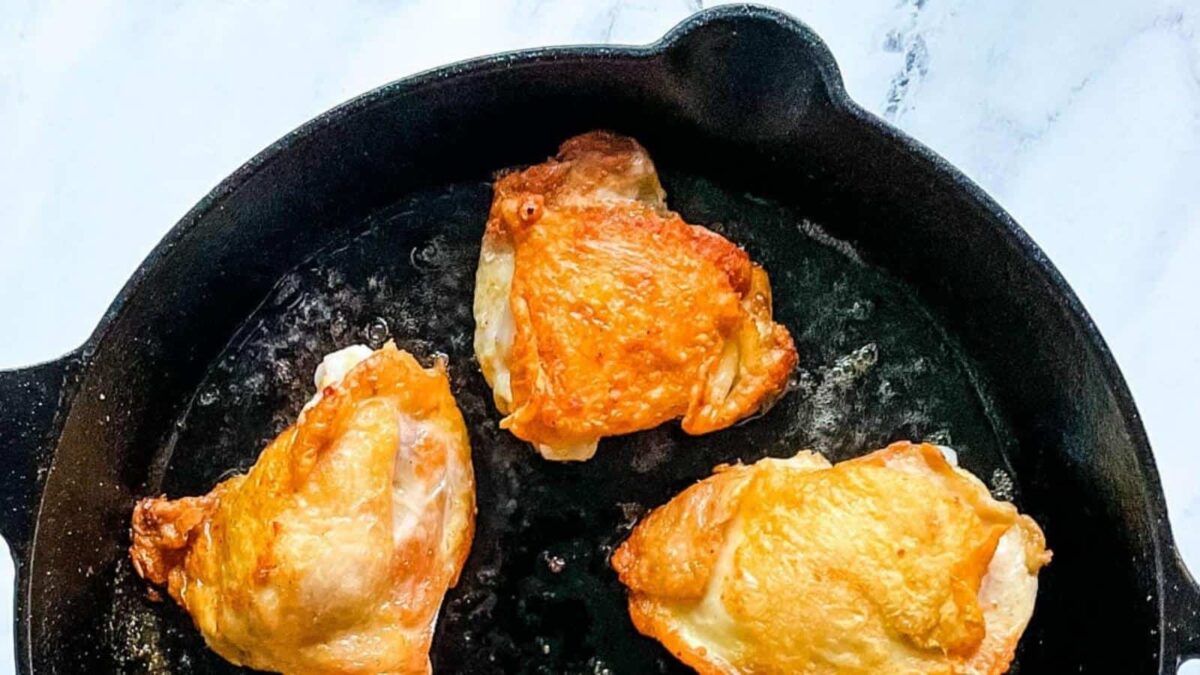 Three pieces of golden-brown chicken thighs with crispy skin cooking in a black cast iron skillet on a white surface—perfect for fans of juicy dishes and classic chicken recipes.