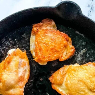 Three pieces of golden-brown chicken thighs with crispy skin cooking in a black cast iron skillet on a white surface—perfect for fans of juicy dishes and classic chicken recipes.
