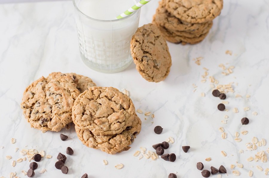 A glass of milk with a striped straw sits next to stacks of healthy cookies—oatmeal chocolate chip vegetarian cookies—surrounded by scattered oats and chocolate chips on a marble surface.