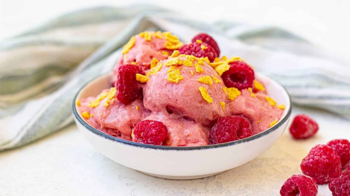 A bowl of pink raspberry sorbet topped with fresh raspberries and cornflake cereal, perfect for fans of yogurt breakfast or those seeking new recipes using yogurt, with a striped cloth in the background.