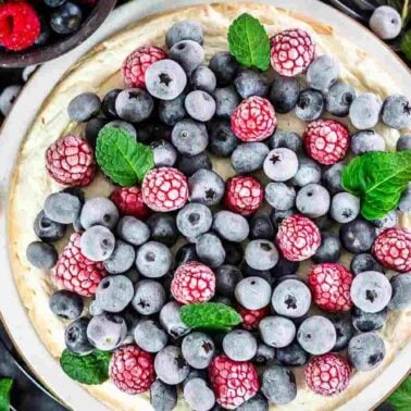 A cheesecake made with creamy yogurt sits on a plate, topped with frozen blueberries, raspberries, and mint leavesโperfect inspiration for your next yogurt recipes adventure. Bowls of berries and fresh mint surround this tempting treat.