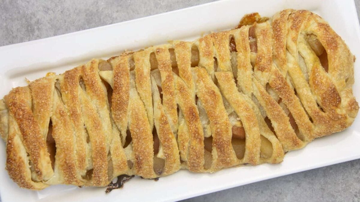 A rectangular apple strudel, one of the classic apple desserts, with a golden, lattice-patterned crust is displayed on a white serving tray in clean cuts and full slices.