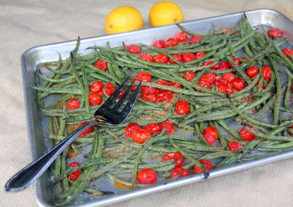 Roasted green beans and cherry tomatoes on a metal baking sheet with a serving fork; two lemons in the background—perfect for fans of easy cooking and delicious sheet pan recipes.