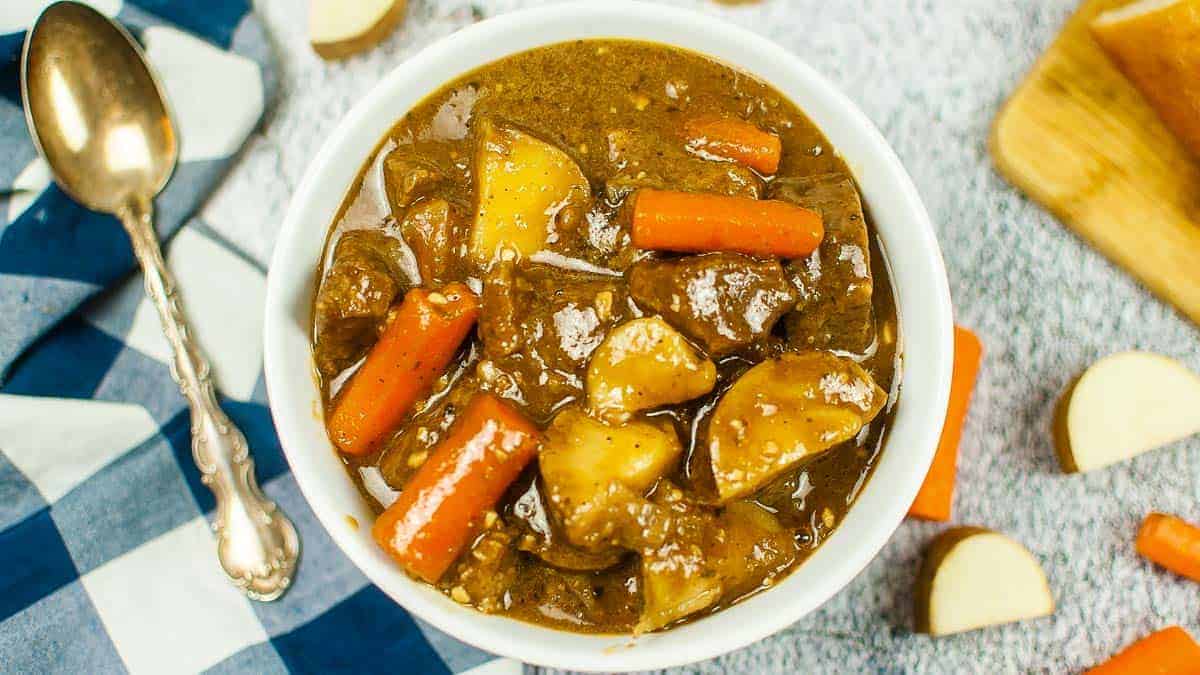 A bowl of beef stew with chunks of beef, potatoes, and carrots in brown gravy—a classic one-pot recipe—sits on a table with a spoon and napkin nearby.