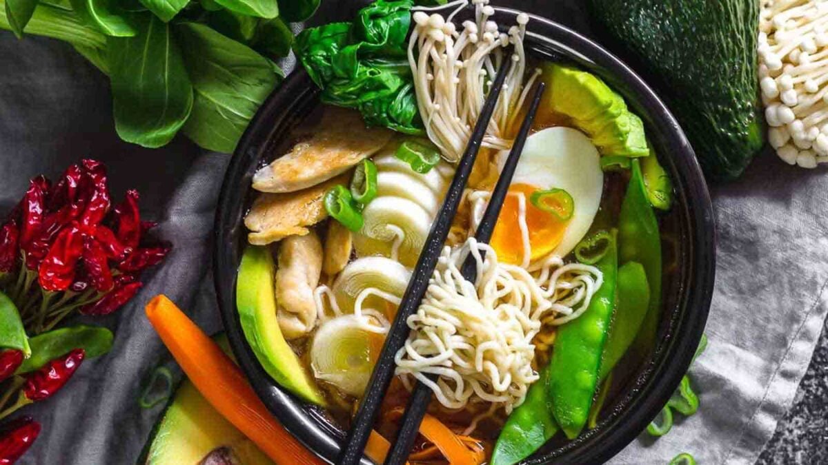 A bowl of ramen with noodles, boiled egg, sliced avocado, mushrooms, and vegetables is surrounded by fresh greens—perfect for those who love flavorful meals and want to simplify cooking with delicious one-pot recipes.