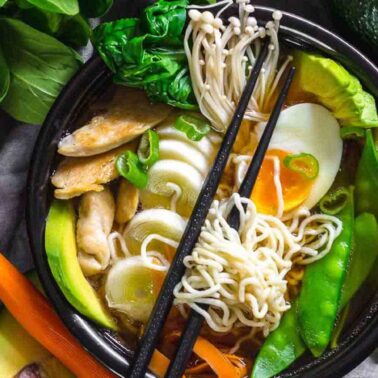 A bowl of ramen with noodles, boiled egg, sliced avocado, mushrooms, and vegetables is surrounded by fresh greensโperfect for those who love flavorful meals and want to simplify cooking with delicious one-pot recipes.