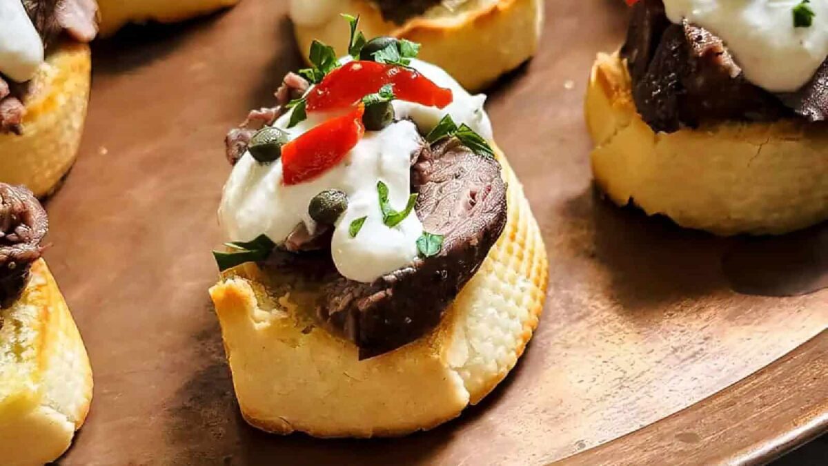Slices of toasted baguette topped with roast beef, creamy sauce, chopped parsley, capers, and red pepper make for creative appetizers, beautifully arranged on a brown plate.