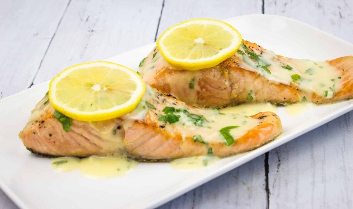 Two cooked salmon fillets topped with lemon slices and a creamy herb sauce are served on a white rectangular plate—an ideal low carb meal for high energy and sustained hunger low.