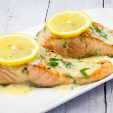 Two cooked salmon fillets topped with lemon slices and a creamy herb sauce are served on a white rectangular plateโan ideal low carb meal for high energy and sustained hunger low.
