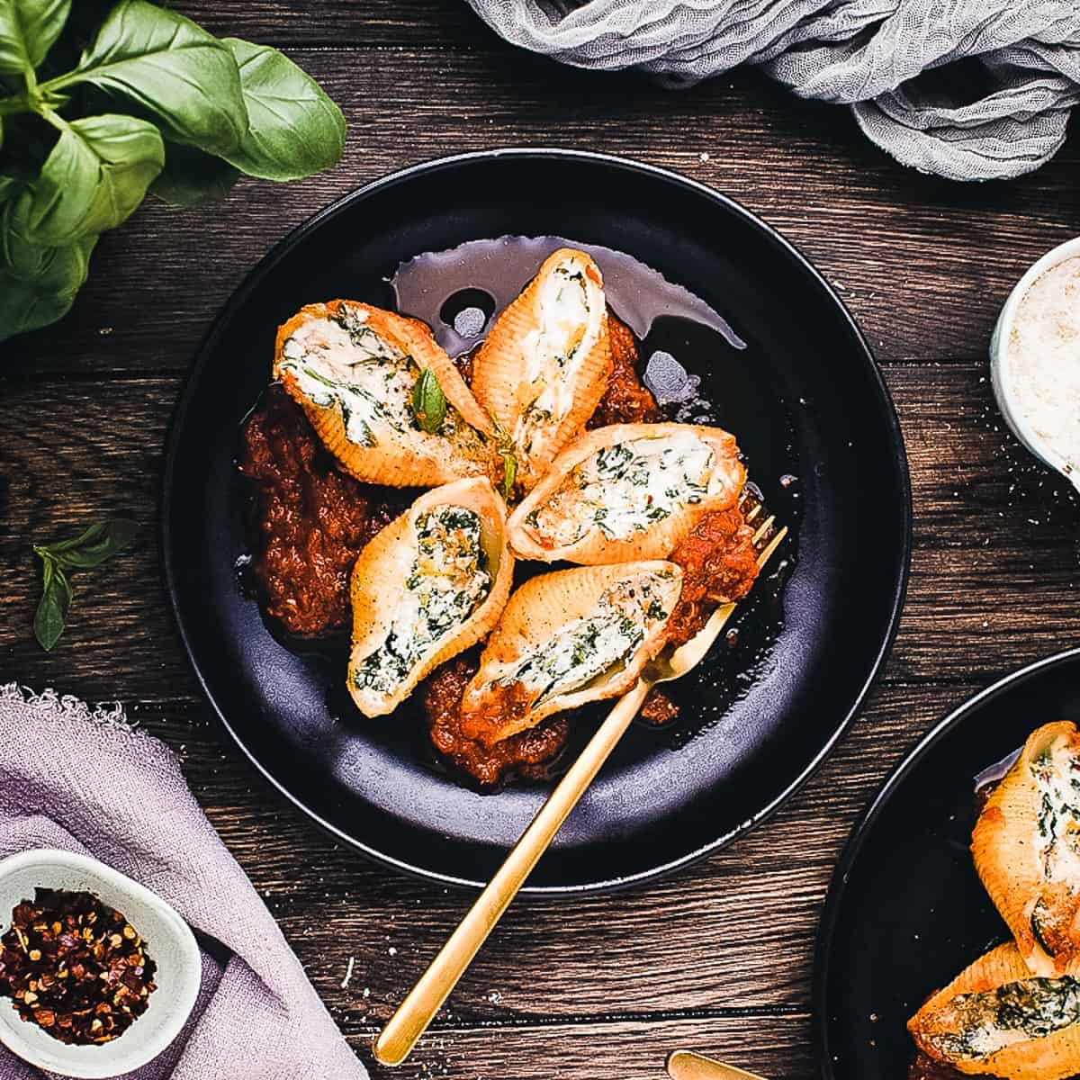 A black plate with large pasta shells stuffed with spinach and creamy ricotta cheese, served on tomato sauce, with a gold fork and garnishes nearby on a wooden table. Perfect for fans of classic ricotta recipes.