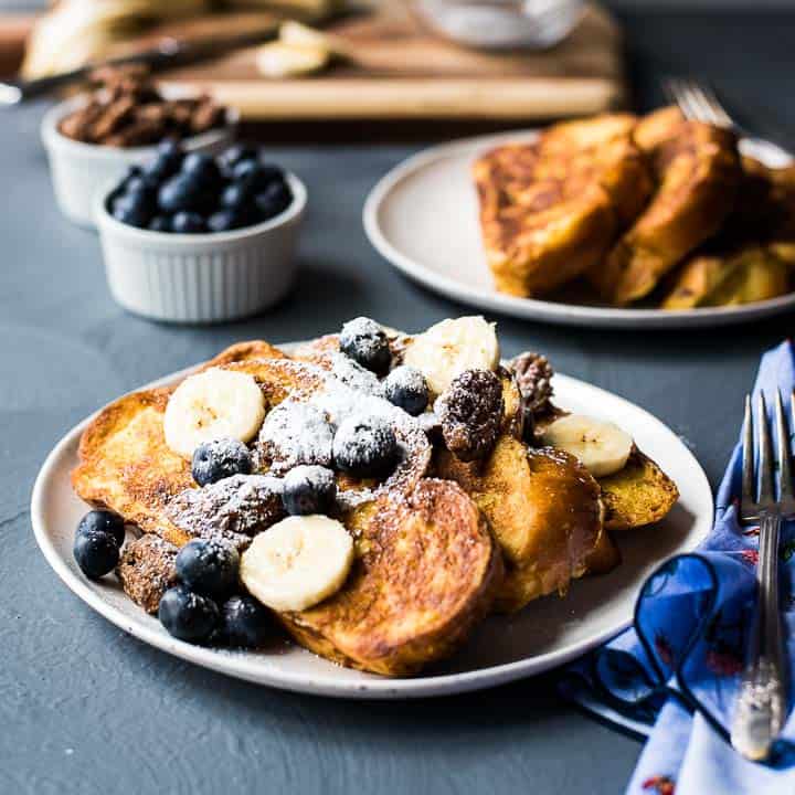 Plate of French toast topped with powdered sugar, banana slices, and blueberries, with a hint of vanilla. A fork sits beside the plate, with extra toast and fruit in the background—perfect for those who love sweet creations.