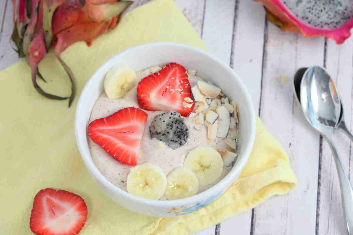 A bowl of smoothie topped with sliced strawberries, banana, coconut flakes, and a piece of dragon fruit on a yellow napkin with a spoon nearby—perfect for kid-friendly recipes that make mealtime fun.