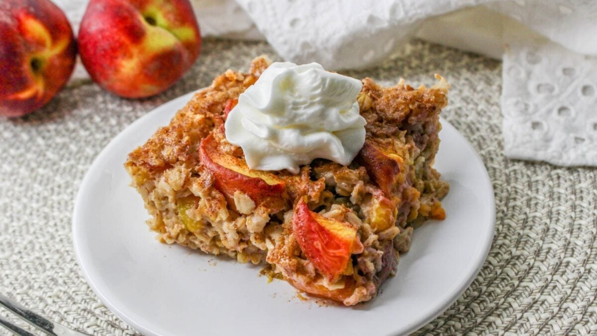 A slice of baked oatmeal with peach slices and a dollop of whipped cream on top, served on a white plate with a peach in the background—perfect for those who love delicious oatmeal recipes.