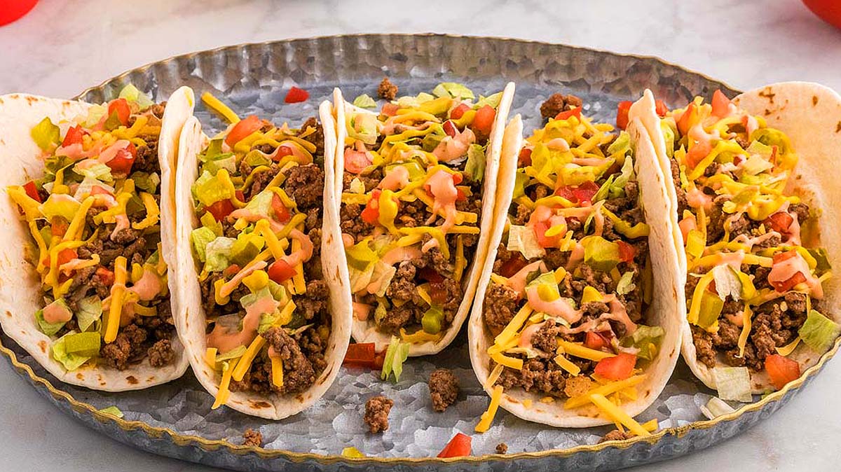 Four soft shell tacos filled with ground beef, shredded cheese, lettuce, diced tomatoes, and a drizzle of sauce are arranged on a metal tray—bringing bold cheeseburger flavor to your table with this creative new dish.
