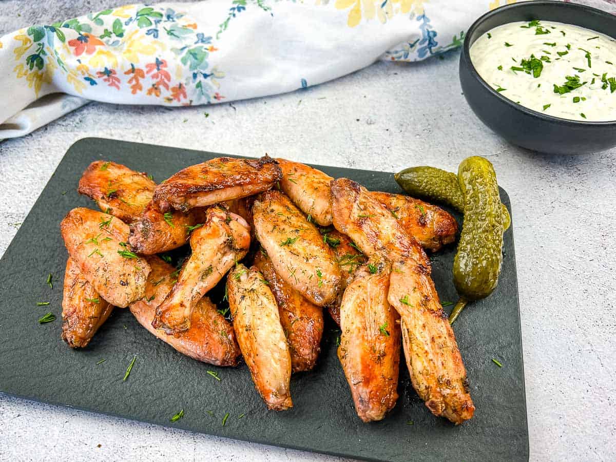 A slate platter with seasoned chicken wings, two pickles, and a bowl of creamy dip garnished with herbs, set on a light surface with a floral napkin in the background—perfect for crowd-pleasing appetizers or game day snacks.