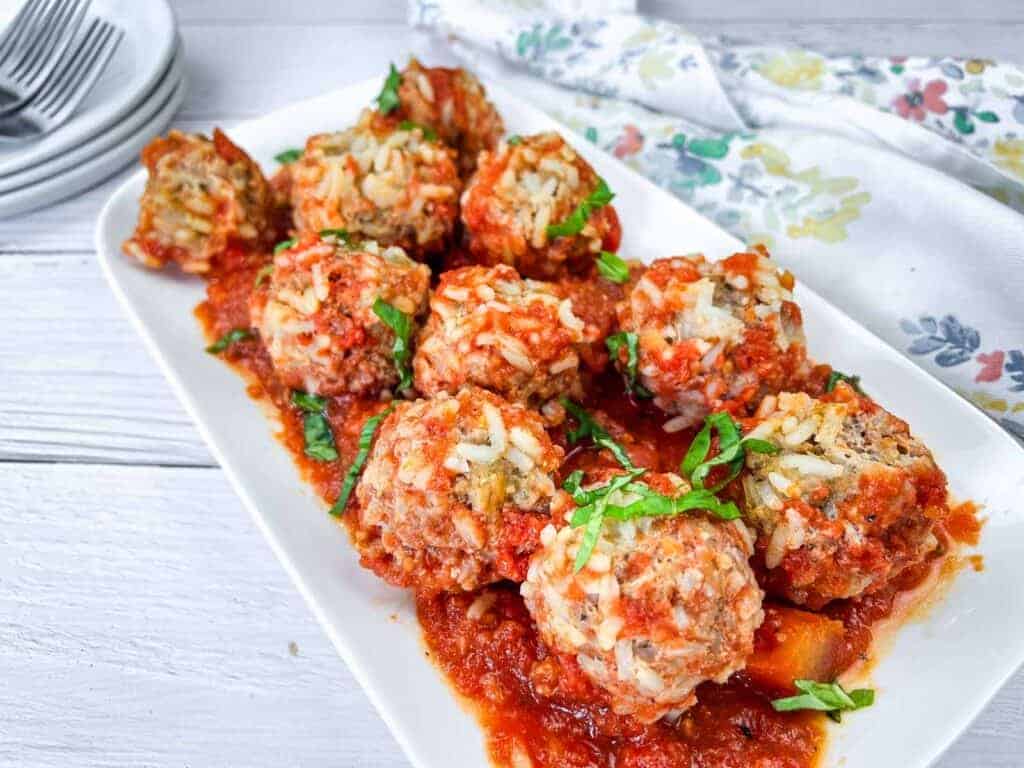 A rectangular white plate with rice-filled meatballs in red tomato sauce, garnished with chopped herbs, sits on a white wooden table beside a floral napkin and stacked plates—the perfect choice for easy meals.