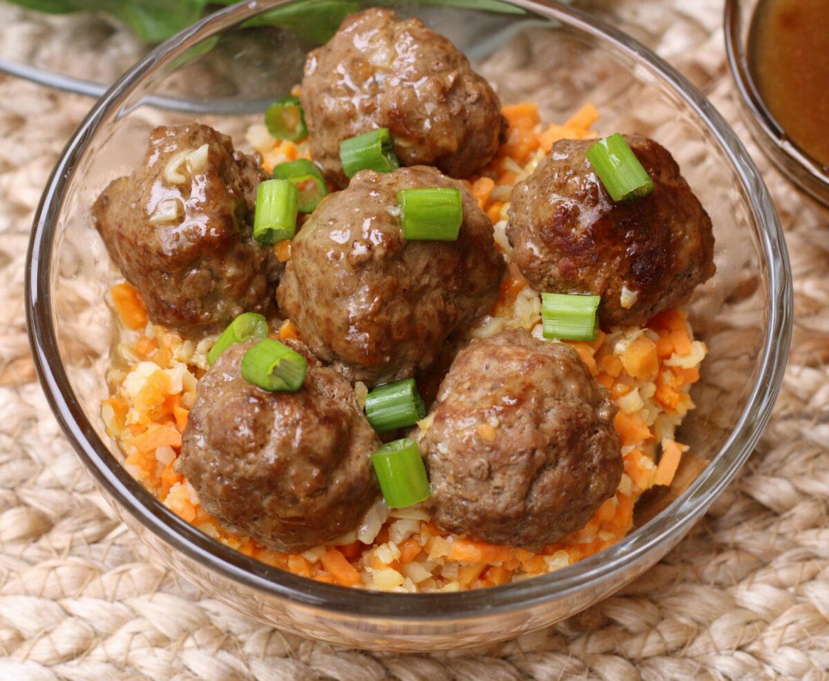 Six cooked meatballs topped with chopped green onions are served over a bed of finely diced carrots and rice in a clear glass bowl—one of our favorite easy meals for quick, fun recipes any night.