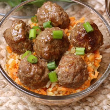 Six cooked meatballs topped with chopped green onions are served over a bed of finely diced carrots and rice in a clear glass bowl—one of our favorite easy meals for quick, fun recipes any night.