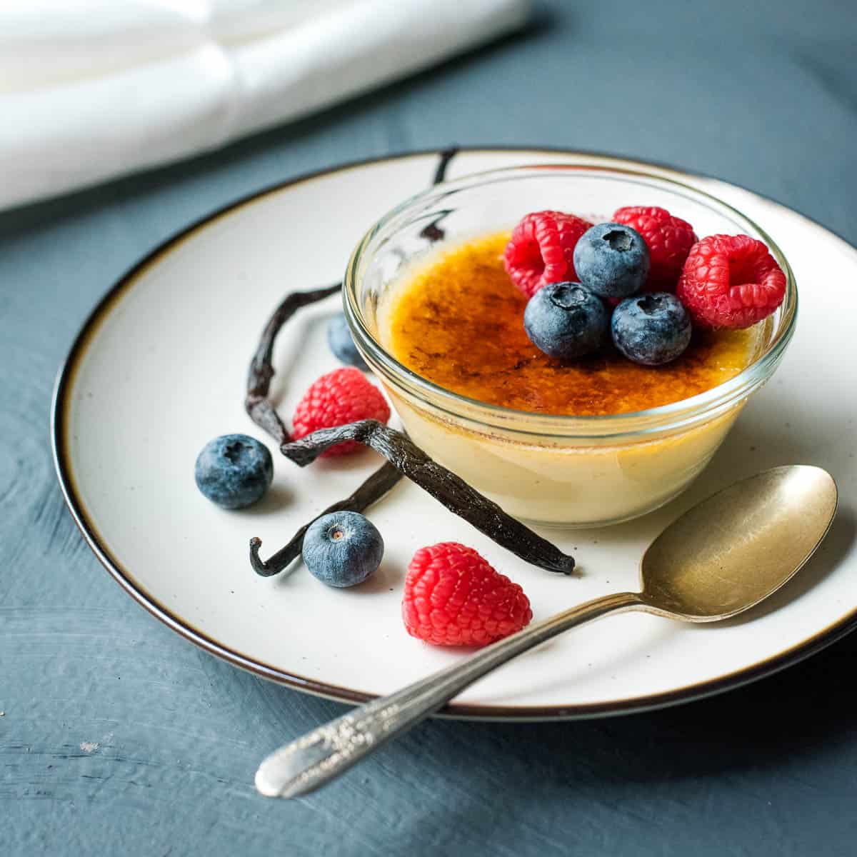 A small glass bowl of crème brûlée, one of the finest vanilla desserts, topped with blueberries and raspberries, served on a white plate with a gold spoon, vanilla beans, and extra berries.