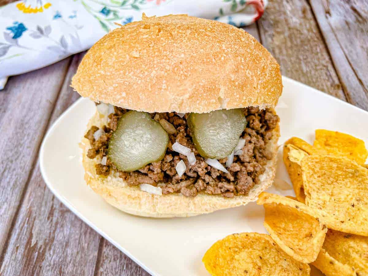 A loose meat sandwich with ground beef, chopped onions, and pickle slices on a bun makes for one of the most unique sandwiches, served on a plate with corn chips.