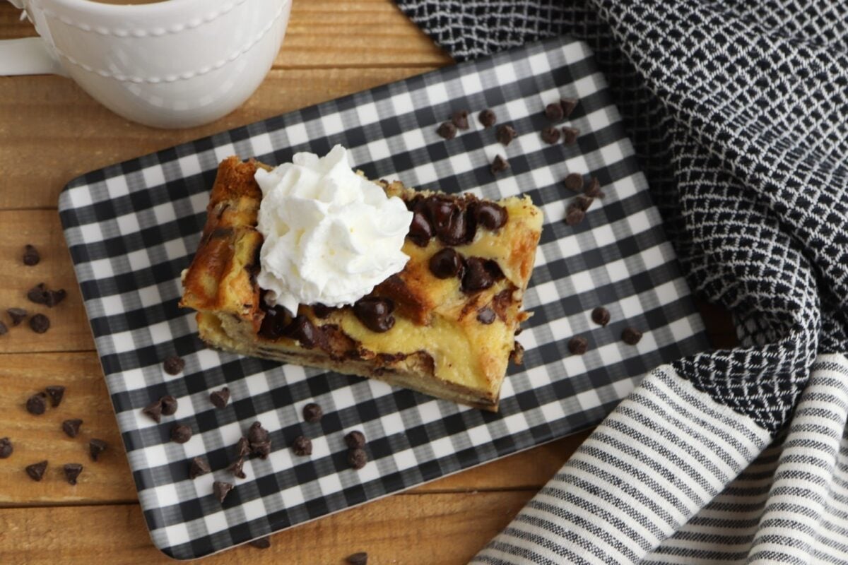 A slice of chocolate chip bread pudding, perfect for simple breakfast recipes, is topped with whipped cream on a black-and-white checkered tray, surrounded by chocolate chips and a striped napkin.