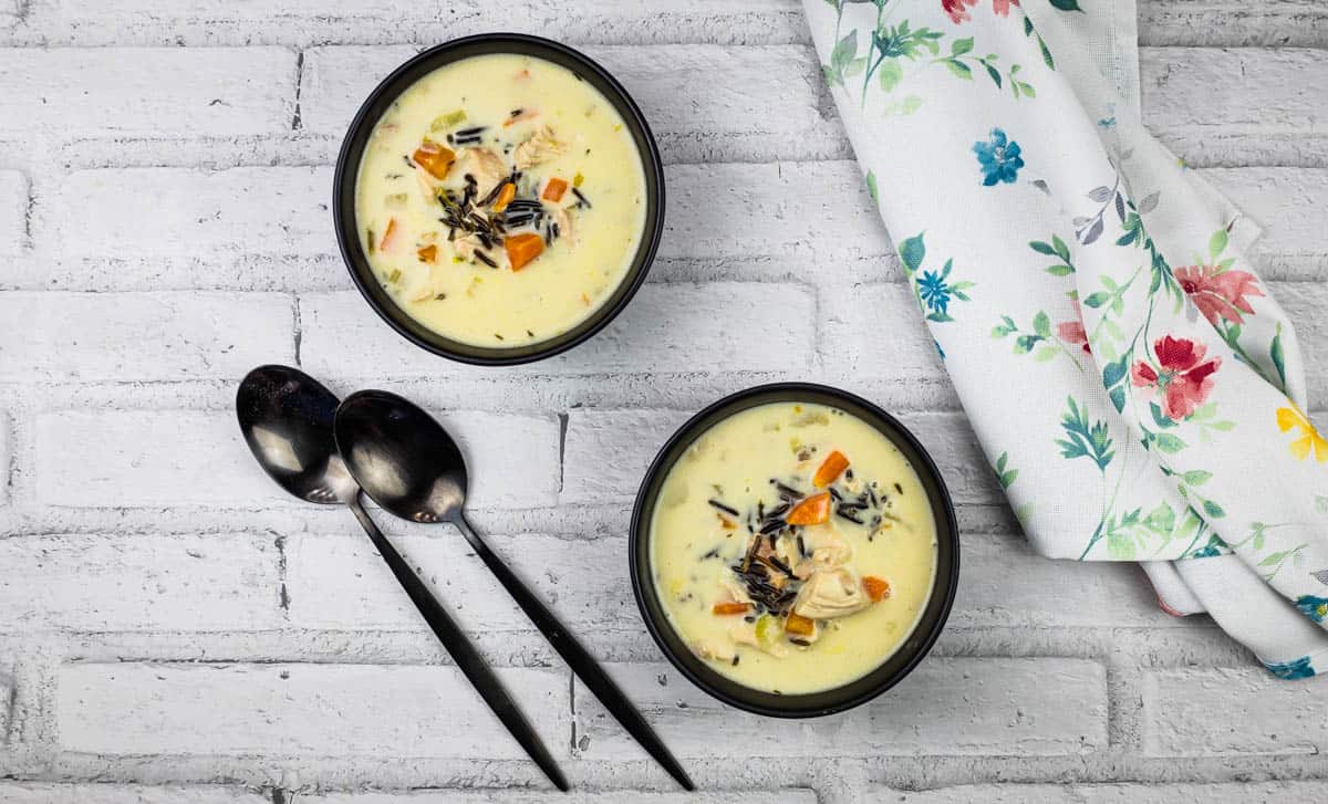 Two bowls of creamy vegetable and wild rice soup, a perfect choice for hearty soups and winter soups, sit on a white brick surface with two black spoons and a floral napkin beside them.