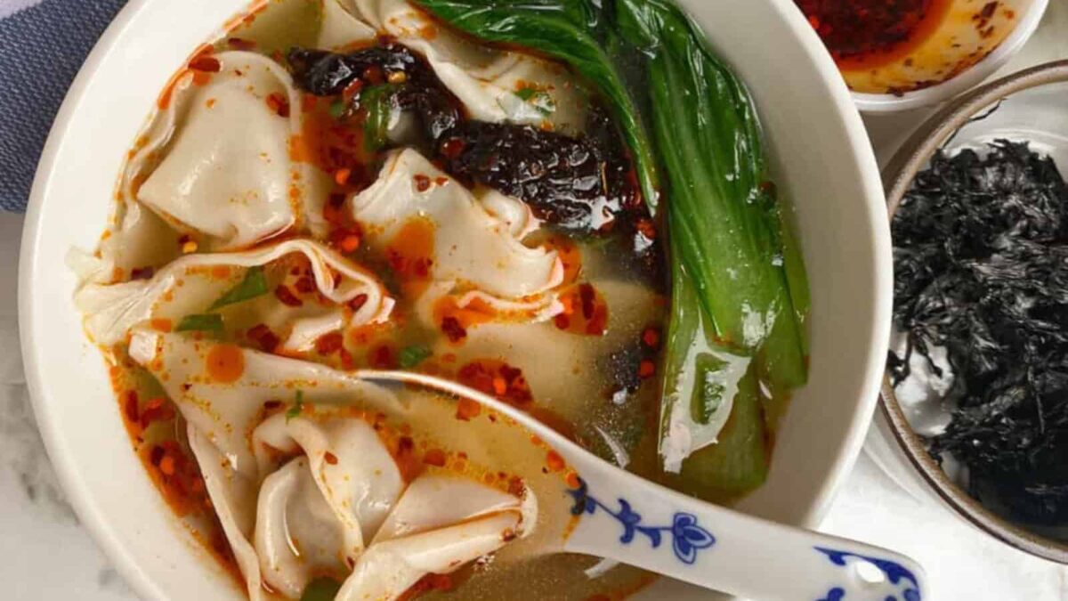 A bowl of hearty soup with wide noodles, leafy greens, and chili oil, served with a ceramic spoon; seaweed and extra chili oil are in small dishes on the side—perfect for comforting winter soups.
