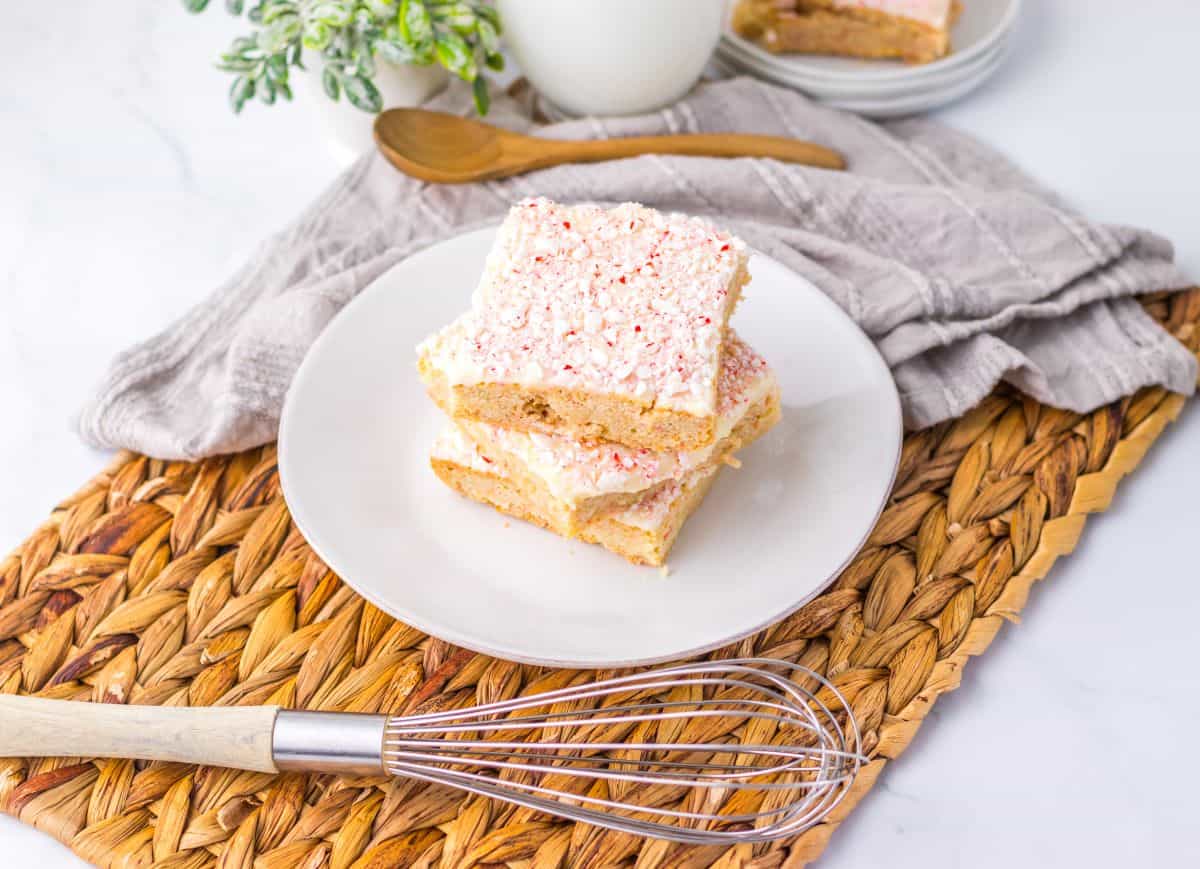 A plate with three frosted dessert bars stacked on top, placed on a woven mat beside a whisk, a gray napkin, and a wooden spoon—perfect for showcasing your favorite peppermint recipes.