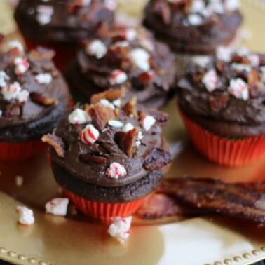 Chocolate cupcakes with chocolate frosting, topped with crumbled bacon and crushed peppermint, arranged on a gold plate with bacon strips—a bold twist on holiday recipes and a fun spin on peppermint desserts.