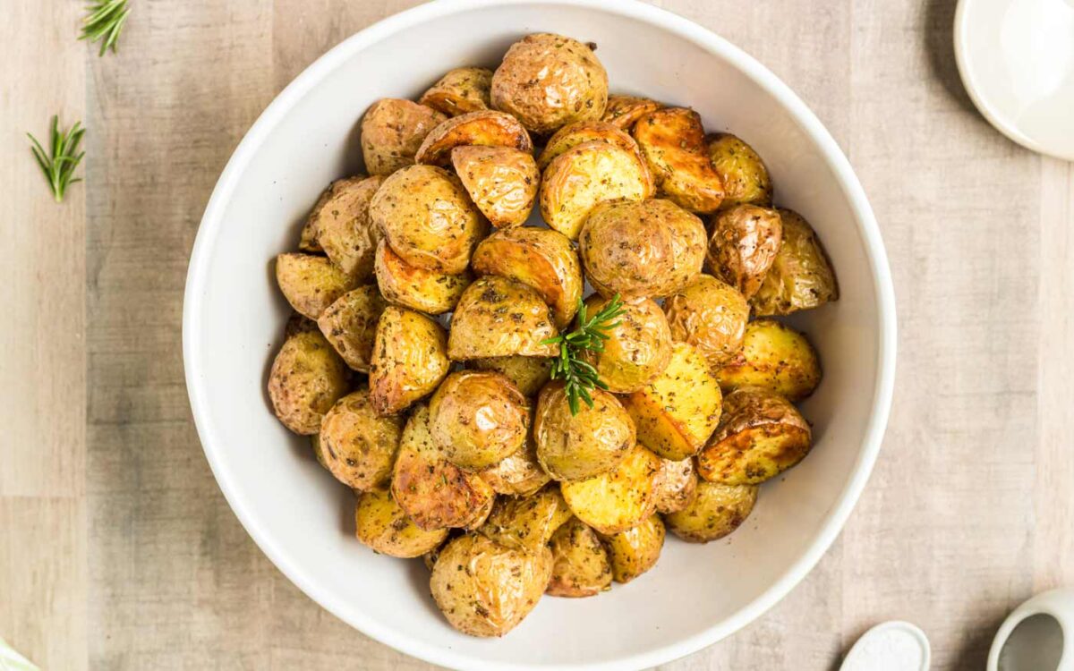 A white bowl filled with roasted baby potatoes, seasoned with herbs, sits on a light-colored table. A small sprig of rosemary garnishes these tasty potatoes—perfect as one of your go-to rosemary sides.