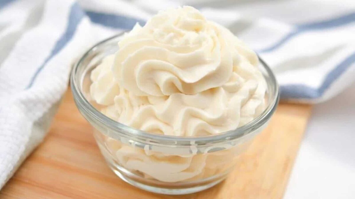 A glass bowl filled with piped swirls of white whipped cream sits on a wooden surface next to a striped cloth, perfect for topping your favorite vanilla desserts or inspiring new sweet creations.