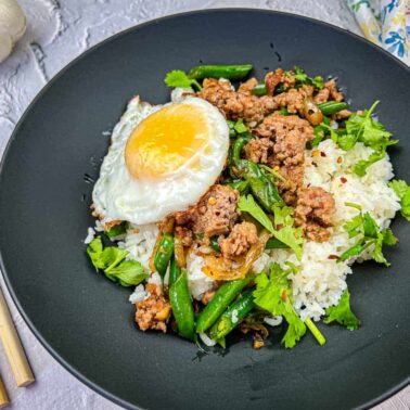 A black plate with white rice, ground meat, green beans, fresh herbs, and a sunny-side-up egg, served with chopsticks on the sideโperfect for those who enjoy Asian-inspired dinners.
