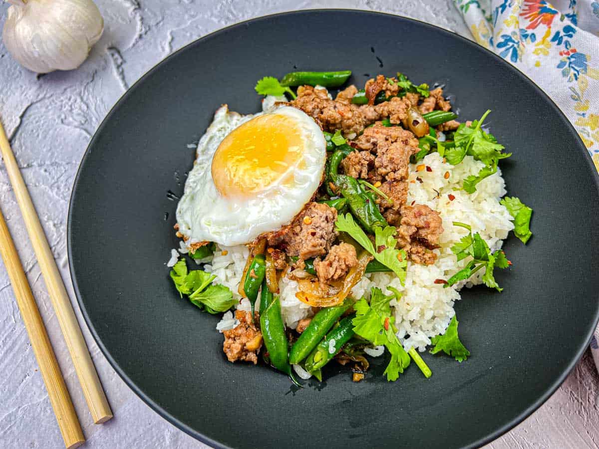 A black plate with white rice, ground meat, green beans, fresh herbs, and a sunny-side-up egg, served with chopsticks on the side—perfect for those who enjoy Asian-inspired dinners.