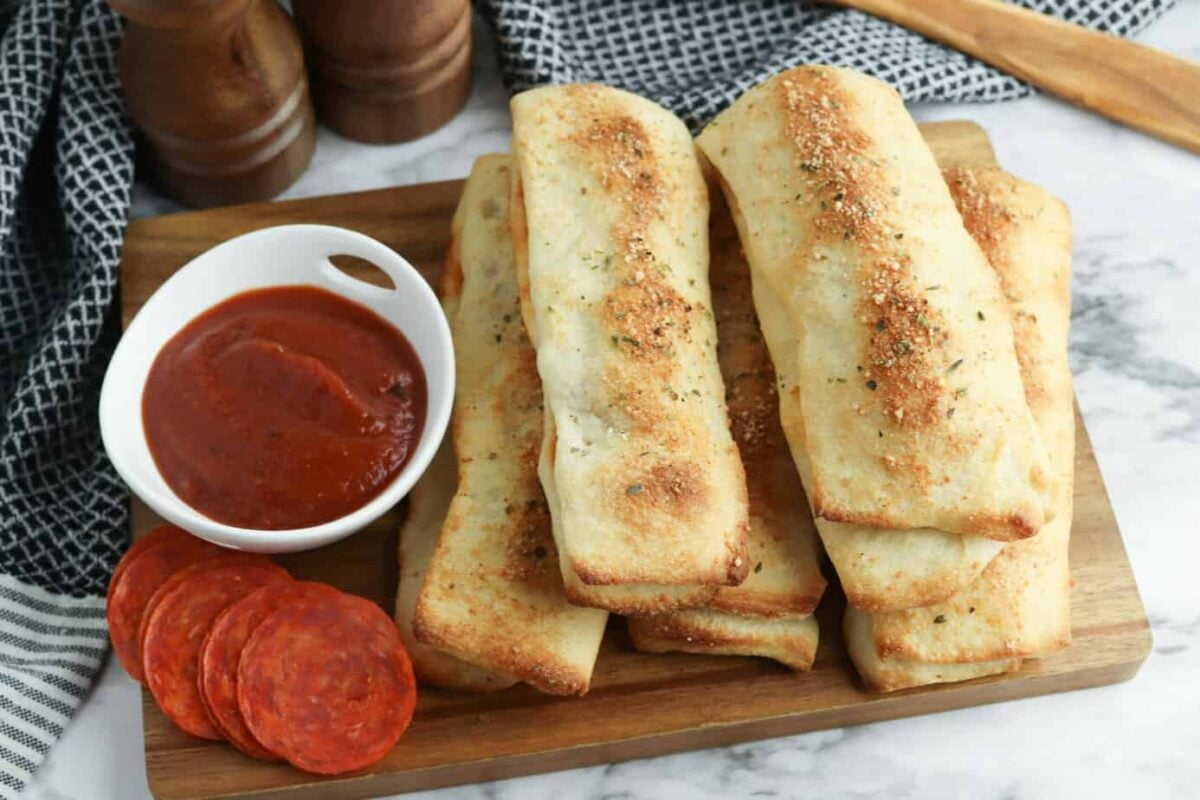A wooden board with several baked breadsticks, a small bowl of marinara sauce, and slices of pepperoni on a marble surface—perfect for air fryer appetizers or other easy appetizer recipes.
