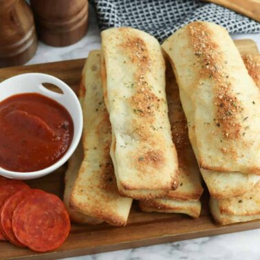 A wooden board with several baked breadsticks, a small bowl of marinara sauce, and slices of pepperoni on a marble surfaceโperfect for air fryer appetizers or other easy appetizer recipes.