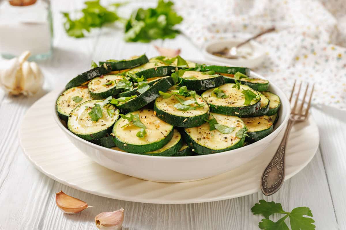 A white bowl filled with sliced, seasoned zucchini—an adaptable ingredient—garnished with chopped herbs, sits on a white plate with a fork beside it on a light wooden table.