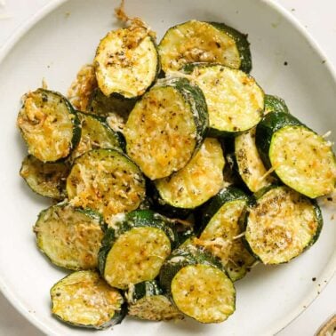 A white plate filled with sliced, roasted zucchiniโan adaptable ingredientโtopped with melted cheese and herbs.